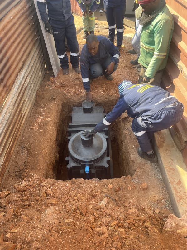 Two installation workers lowering the black Bio-Separator chamber into an excavated trench beside a home, with the crew looking on