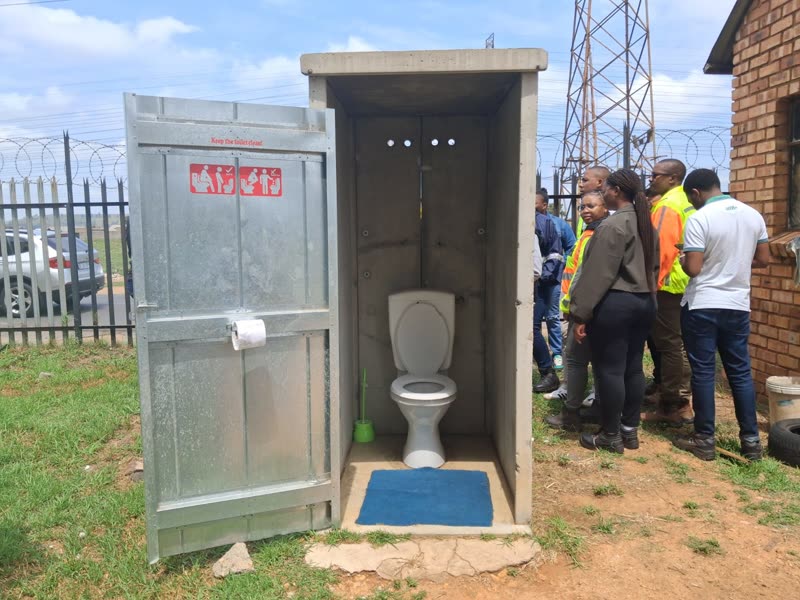 A completed unit with its galvanised door open, showing the flush toilet, brush and mat inside