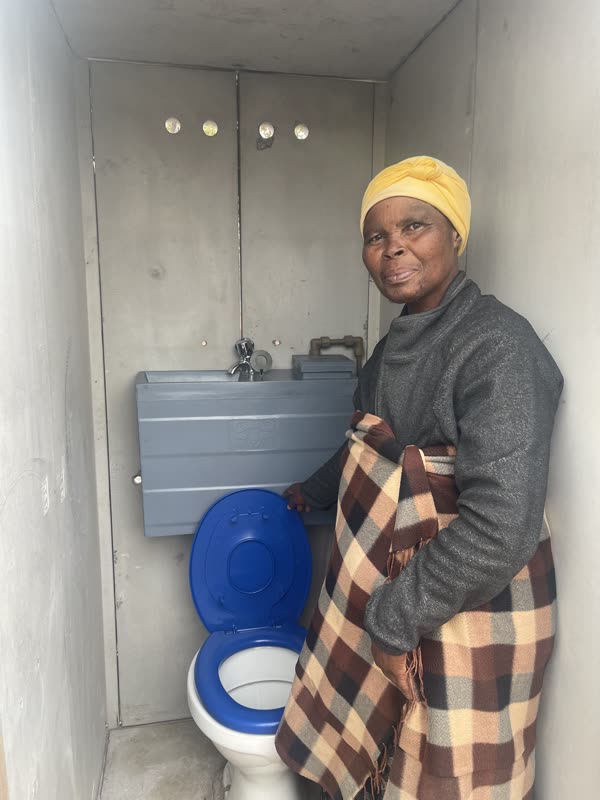 A resident standing beside the flush toilet and hand-wash basin inside her completed unit